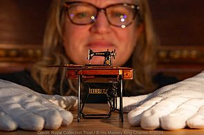 Miniatur-Nähmaschine der Firma Singer, im Hintergrund das Gesicht einer Frau, die ihre Hände neben die Nähmaschine gelegt hat